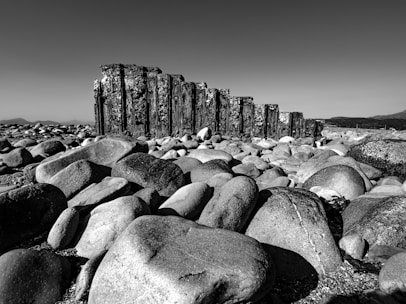 A landscape featuring a large collection of smooth, rounded stones scattered across the ground. A weathered wooden structure, resembling an old breakwater, stands prominently in the middle distance. The backdrop is a clear sky and faint outlines of mountains, creating a monochromatic, serene atmosphere.