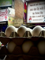 A stack of white eggs is arranged in a cardboard carton against a blurry urban backdrop with signage and buildings. The eggs are the focal point and are well-lit, while the background is out of focus.