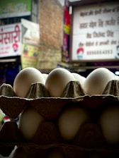 A stack of white eggs is arranged in a cardboard carton against a blurry urban backdrop with signage and buildings. The eggs are the focal point and are well-lit, while the background is out of focus.
