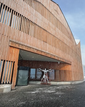 A modern building with a wooden facade featuring vertical slats. A sculpture resembling a human figure is positioned near the entrance, and a few snowflakes are visible in the air. The structure has curved architectural lines and an inscription with Cyrillic characters can be seen above the entrance.
