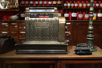 An antique cash register with an ornate design is prominently displayed, featuring a label that reads 'National Dayton, Ohio, U.S.A.' The setting is a vintage store or apothecary with wooden shelves and cabinets in the background, filled with labeled jars of various substances. A green, vintage upright lamp is visible on one side, while an old mechanical calculator is placed on the other side of the cash register.