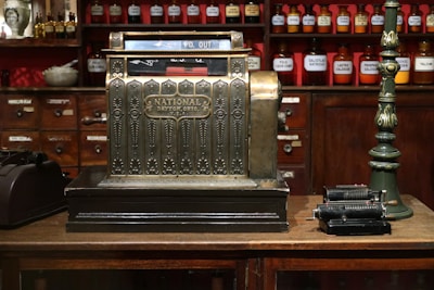 An antique cash register with an ornate design is prominently displayed, featuring a label that reads 'National Dayton, Ohio, U.S.A.' The setting is a vintage store or apothecary with wooden shelves and cabinets in the background, filled with labeled jars of various substances. A green, vintage upright lamp is visible on one side, while an old mechanical calculator is placed on the other side of the cash register.