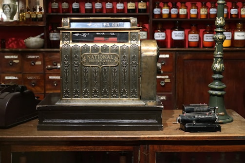 An antique cash register with an ornate design is prominently displayed, featuring a label that reads 'National Dayton, Ohio, U.S.A.' The setting is a vintage store or apothecary with wooden shelves and cabinets in the background, filled with labeled jars of various substances. A green, vintage upright lamp is visible on one side, while an old mechanical calculator is placed on the other side of the cash register.