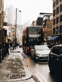 Street scene with a red double-decker bus labeled Shoreditch, 47, driven on a crowded city road. Cars are parked and moving beside the bus, with pedestrians walking on the sidewalk. The buildings lining the street are a blend of modern and older architecture. It's an overcast day with wet pavement, indicating recent rain.