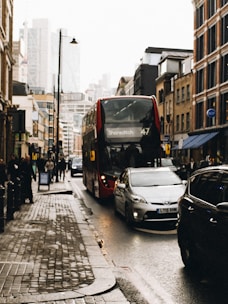 Street scene with a red double-decker bus labeled Shoreditch, 47, driven on a crowded city road. Cars are parked and moving beside the bus, with pedestrians walking on the sidewalk. The buildings lining the street are a blend of modern and older architecture. It's an overcast day with wet pavement, indicating recent rain.