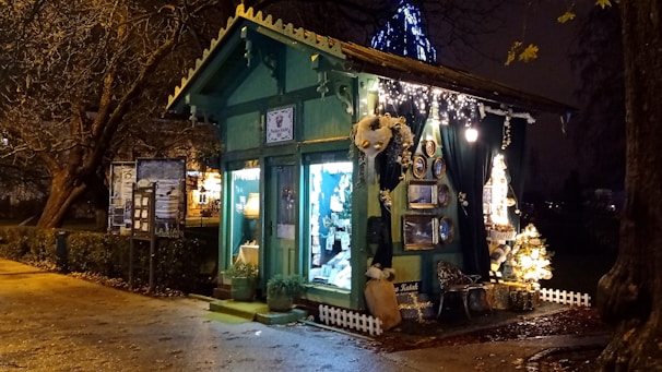 A small, charming wooden shop with a green exterior, adorned with decorative elements and fairy lights that create a cozy and festive atmosphere. The interior is softly illuminated, displaying various items, possibly crafts or gifts, visible through the large window panes. The setting is outdoors at night, surrounded by trees and a gentle glow from streetlights.