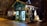 A small, charming wooden shop with a green exterior, adorned with decorative elements and fairy lights that create a cozy and festive atmosphere. The interior is softly illuminated, displaying various items, possibly crafts or gifts, visible through the large window panes. The setting is outdoors at night, surrounded by trees and a gentle glow from streetlights.