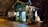 A small, charming wooden shop with a green exterior, adorned with decorative elements and fairy lights that create a cozy and festive atmosphere. The interior is softly illuminated, displaying various items, possibly crafts or gifts, visible through the large window panes. The setting is outdoors at night, surrounded by trees and a gentle glow from streetlights.