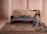 A woman in traditional attire is seated on a vintage-style wooden and cane sofa with gray cushions. The setting features a richly patterned carpet and a neutral-toned background with classical columns.