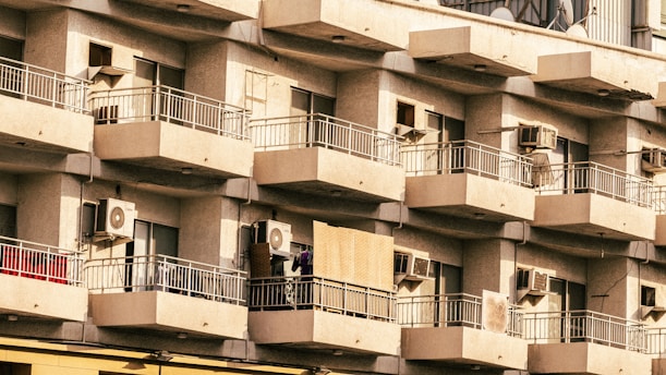 A residential building with multiple balconies featuring air conditioning units on each one. The balconies have metal railings, and some show signs of everyday life such as clothes and other items. The building's facade is uniform and has a somewhat aged appearance.