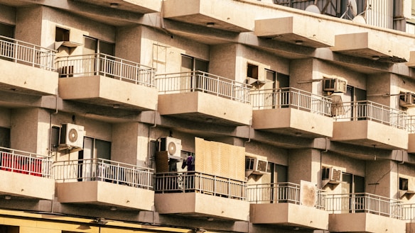 A residential building with multiple balconies featuring air conditioning units on each one. The balconies have metal railings, and some show signs of everyday life such as clothes and other items. The building's facade is uniform and has a somewhat aged appearance.