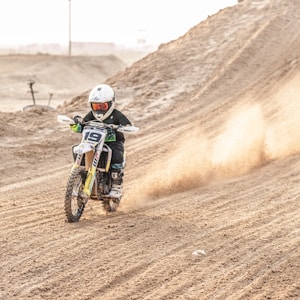 A motorcyclist wearing protective gear and a helmet speeds across a sandy terrain, leaving a trail of dust behind. The background shows the arid landscape of the desert with smooth sand dunes.