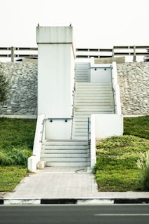A set of white concrete stairs with railings leads up to a flat platform. The structure is set against a background of a stone wall and barrier, with green grass on either side. The sky is light in color, and there are a few pigeons perched on top of the structure.