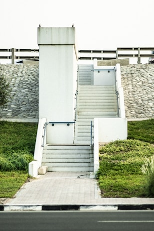 A set of white concrete stairs with railings leads up to a flat platform. The structure is set against a background of a stone wall and barrier, with green grass on either side. The sky is light in color, and there are a few pigeons perched on top of the structure.