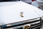 A close-up view of the front of a white car, featuring the logo of a well-known automobile manufacturer. The hood ornament is prominently displayed in gold, situated above the chrome grill of the vehicle.
