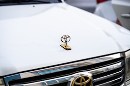 A close-up view of the front of a white car, featuring the logo of a well-known automobile manufacturer. The hood ornament is prominently displayed in gold, situated above the chrome grill of the vehicle.