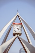 A large, modern clock structure with an Omega logo on a red background is prominently displayed. The clock is encircled by a geometric framework of white supports converging at the top, forming a triangular shape. In the background, a building with another clock at the top is visible against a clear blue sky.