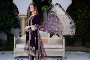 A woman is standing in an outdoor setting wearing a traditional dark purple outfit with intricate embellishments. She has a sheer dupatta with a combination of white and purple colors and detailed embroidery. She is posing beside an antique-style white bench with intricate carvings. The background includes lush green foliage and classical architectural elements.