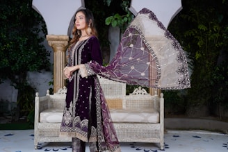 A woman is standing in an outdoor setting wearing a traditional dark purple outfit with intricate embellishments. She has a sheer dupatta with a combination of white and purple colors and detailed embroidery. She is posing beside an antique-style white bench with intricate carvings. The background includes lush green foliage and classical architectural elements.