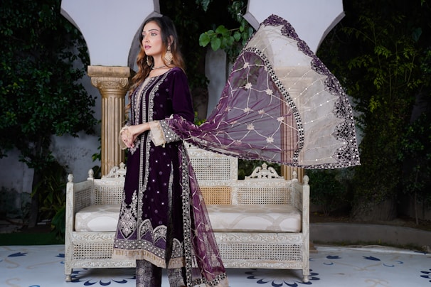 A woman is standing in an outdoor setting wearing a traditional dark purple outfit with intricate embellishments. She has a sheer dupatta with a combination of white and purple colors and detailed embroidery. She is posing beside an antique-style white bench with intricate carvings. The background includes lush green foliage and classical architectural elements.