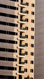 Tall residential building with a series of evenly spaced balconies featuring green railings. The facade is composed of beige concrete, with rows of rectangular windows. Shadows create a strong contrast on the surface of the building.