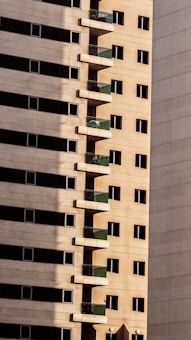 Tall residential building with a series of evenly spaced balconies featuring green railings. The facade is composed of beige concrete, with rows of rectangular windows. Shadows create a strong contrast on the surface of the building.