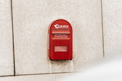 A bright red Emirates Post letter mailbox is mounted on a textured, off-white concrete wall. The surface of the wall is slightly stained near the bottom of the mailbox. Arabic and English text are clearly visible on the mailbox, along with the Emirates Post logo.