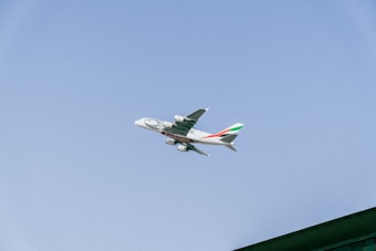 A large commercial airplane with branding on the fuselage flies through a clear blue sky. The aircraft has distinct markings and colorful tail design, suggesting it's an international airline. In the bottom right corner, a structure with a green roof is partially visible.