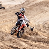 A motorcyclist wearing protective gear rides a dirt bike across a sandy, uneven track. The rider is navigating a turn, kicking up sand behind the bike. The environment is rugged and the rider appears focused and in control, indicating a high level of skill in handling the bike.