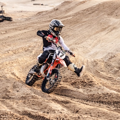 A motorcyclist wearing protective gear rides a dirt bike across a sandy, uneven track. The rider is navigating a turn, kicking up sand behind the bike. The environment is rugged and the rider appears focused and in control, indicating a high level of skill in handling the bike.