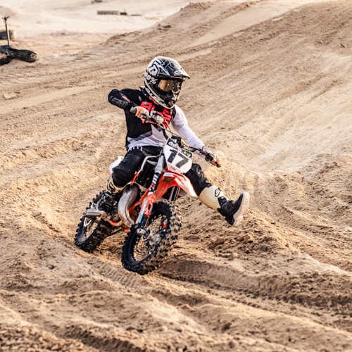 A motorcyclist wearing protective gear rides a dirt bike across a sandy, uneven track. The rider is navigating a turn, kicking up sand behind the bike. The environment is rugged and the rider appears focused and in control, indicating a high level of skill in handling the bike.