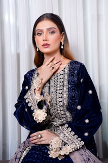 A woman is elegantly dressed in a richly embroidered navy blue outfit with intricate gold detailing. She is wearing matching jewelry, including earrings, rings, and a necklace, and her makeup is tastefully applied. The backdrop consists of soft white curtains.