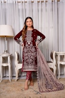 A woman stands elegantly wearing a maroon embroidered traditional dress with intricate detailing. She is positioned indoors, flanked by two chairs and a lamp, against a backdrop of sheer curtains.