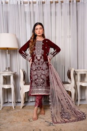 A woman stands elegantly wearing a maroon embroidered traditional dress with intricate detailing. She is positioned indoors, flanked by two chairs and a lamp, against a backdrop of sheer curtains.
