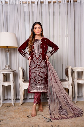 A woman stands elegantly wearing a maroon embroidered traditional dress with intricate detailing. She is positioned indoors, flanked by two chairs and a lamp, against a backdrop of sheer curtains.
