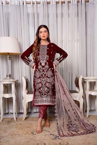 A woman stands elegantly wearing a maroon embroidered traditional dress with intricate detailing. She is positioned indoors, flanked by two chairs and a lamp, against a backdrop of sheer curtains.