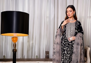 A woman stands elegantly in a detailed embroidered outfit featuring intricate floral patterns. She accessorizes with a necklace, bracelet, earrings, and a ring, highlighting a sophisticated fashion sense. Beside her is a modern lamp with a black shade, contrasting against flowing white curtains in the background.