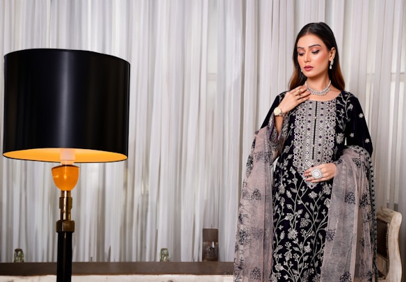 A woman stands elegantly in a detailed embroidered outfit featuring intricate floral patterns. She accessorizes with a necklace, bracelet, earrings, and a ring, highlighting a sophisticated fashion sense. Beside her is a modern lamp with a black shade, contrasting against flowing white curtains in the background.