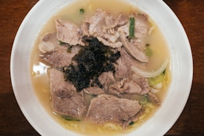 A bowl filled with a light-colored broth containing slices of cooked meat, green onions, and seaweed. The dish is presented in a white bowl placed on a wooden surface.
