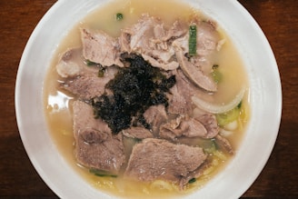 A bowl filled with a light-colored broth containing slices of cooked meat, green onions, and seaweed. The dish is presented in a white bowl placed on a wooden surface.