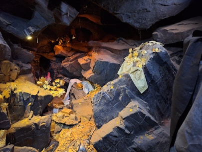 A detailed underground cavern scene with large, jagged rock formations. Scattered throughout the scene are small gold coins and what appear to be golden leaves or decorations. There are areas with a warm, orange lighting that adds depth and a mysterious atmosphere.