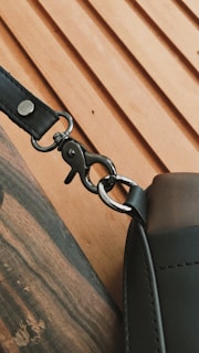 A close-up view of a metal clasp attaching a strap to a leather bag. The clasp is shiny and well-defined, while the leather appears smooth and slightly textured. The background features a light wood surface with diagonal grooves.