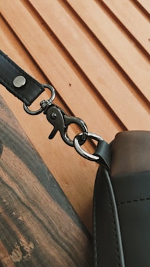 A close-up view of a metal clasp attaching a strap to a leather bag. The clasp is shiny and well-defined, while the leather appears smooth and slightly textured. The background features a light wood surface with diagonal grooves.