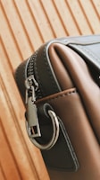 A close-up of a stylish leather bag with a metal zipper and a sturdy metal ring on its side. The bag's material appears smooth and high-quality, with careful stitching visible along the edges. The background features a wooden pattern with vertical lines.
