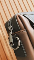 A close-up of a stylish leather bag with a metal zipper and a sturdy metal ring on its side. The bag's material appears smooth and high-quality, with careful stitching visible along the edges. The background features a wooden pattern with vertical lines.