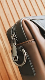 A close-up of a stylish leather bag with a metal zipper and a sturdy metal ring on its side. The bag's material appears smooth and high-quality, with careful stitching visible along the edges. The background features a wooden pattern with vertical lines.