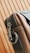 A close-up of a stylish leather bag with a metal zipper and a sturdy metal ring on its side. The bag's material appears smooth and high-quality, with careful stitching visible along the edges. The background features a wooden pattern with vertical lines.