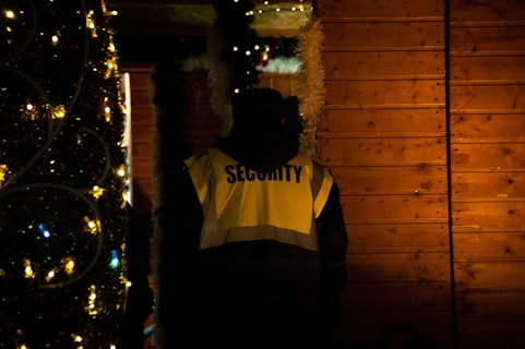 A person wearing a high-visibility jacket with the word 'Security' is standing near a wooden wall adorned with festive decorations, including lights and garlands. The scene is dimly lit, creating a warm, cozy atmosphere.