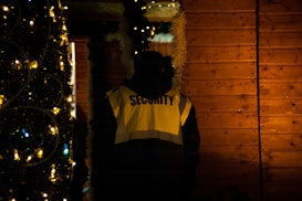 A person wearing a high-visibility jacket with the word 'Security' is standing near a wooden wall adorned with festive decorations, including lights and garlands. The scene is dimly lit, creating a warm, cozy atmosphere.