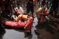 A group of people participate in a ritual involving prostration and prayer in a wet environment. They are wearing colorful traditional clothing with some individuals fully lying on the ground while a woman nearby is in a prayer position. The water on the ground suggests a sacred or ceremonial setting.